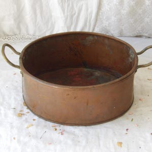 May include: A round copper basin featuring two curved metal handles on opposite sides. The exterior and interior surfaces display a weathered, oxidised patina with visible dark spots and green verdigris, showing a deep metallic brown colour.