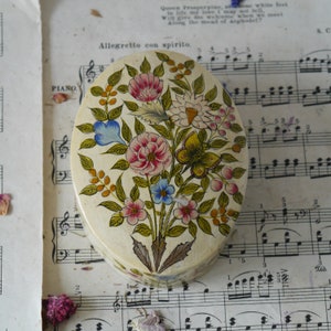 May include: An oval-shaped trinket box with a floral design. The box is painted with a white background and features a variety of colorful flowers, including pink, blue, yellow, and green. The flowers are arranged in a bouquet-like pattern and are surrounded by green leaves. The box is decorated with a butterfly in the center.