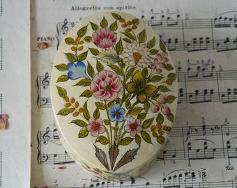 May include: An oval-shaped trinket box with a floral design. The box is painted with a white background and features a variety of colorful flowers, including pink, blue, yellow, and green. The flowers are arranged in a bouquet-like pattern and are surrounded by green leaves. The box is decorated with a butterfly in the center.