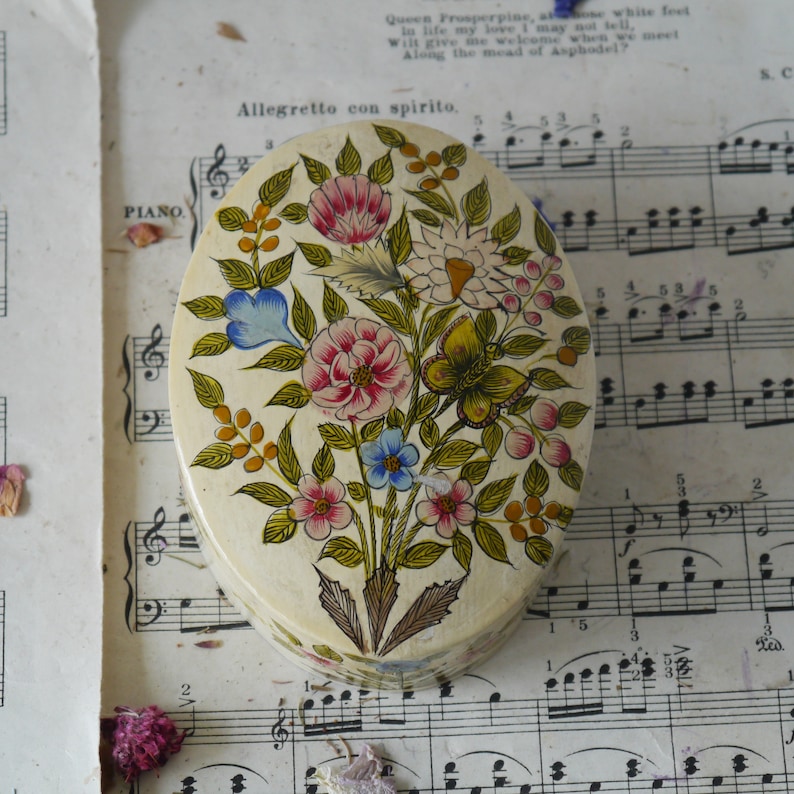 May include: An oval-shaped trinket box with a floral design. The box is painted with a white background and features a variety of colorful flowers, including pink, blue, yellow, and green. The flowers are arranged in a bouquet-like pattern and are surrounded by green leaves. The box is decorated with a butterfly in the center.