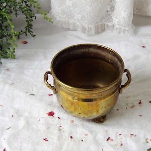 Könnte beinhalten: Eine kleine, antike Messingschale mit zwei verzierten Griffen und drei kleinen Füßen. Die Schale hat eine runde Form und eine warme, goldene Farbe, die Gebrauchsspuren aufweist. Das Innere ist dunkler, möglicherweise durch Nutzung.