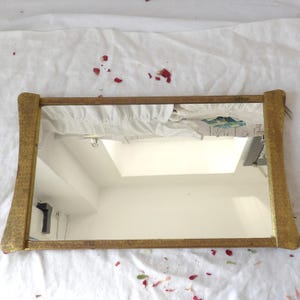 May include: A rectangular mirror with a gold-colored frame. The frame has a textured pattern and curved edges. The mirror reflects a white ceiling and a portion of a room.