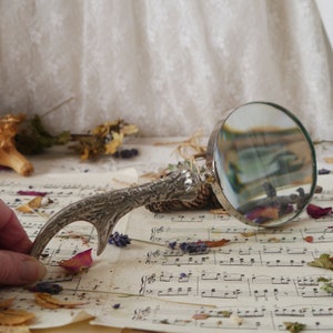 Peut inclure: Une loupe argent&eacute;e avec une poign&eacute;e d&eacute;corative en forme de bois de cerf. La loupe est pos&eacute;e sur une feuille de musique avec des fleurs et des feuilles s&eacute;ch&eacute;es &eacute;parpill&eacute;es autour.