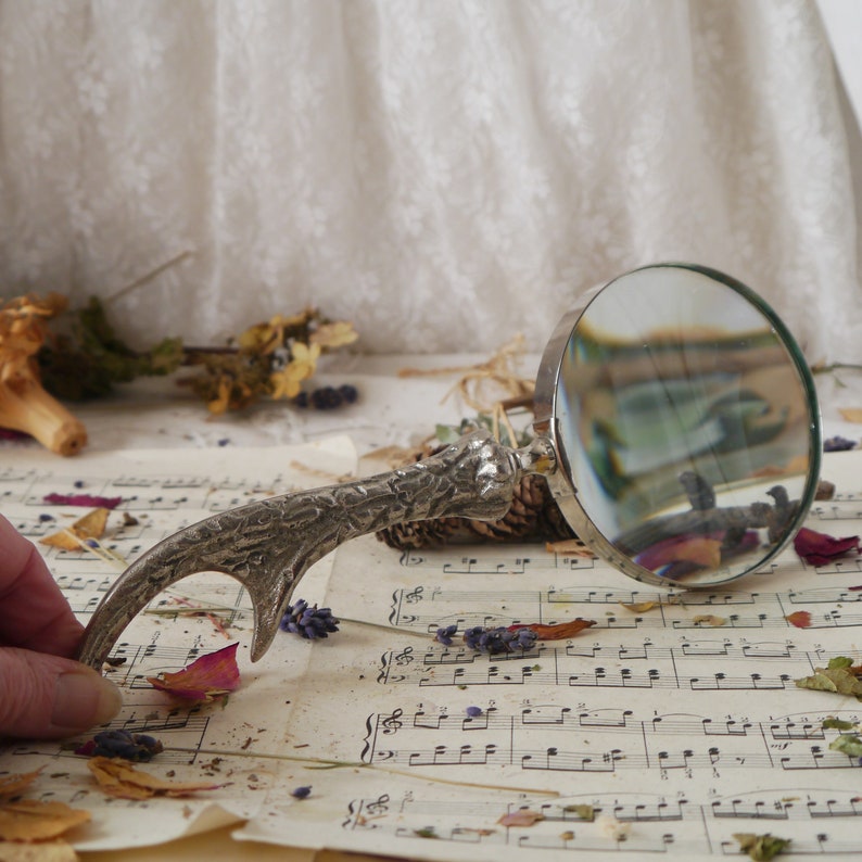 Peut inclure: Une loupe argent&eacute;e avec une poign&eacute;e d&eacute;corative en forme de bois de cerf. La loupe est pos&eacute;e sur une feuille de musique avec des fleurs et des feuilles s&eacute;ch&eacute;es &eacute;parpill&eacute;es autour.