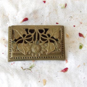 May include: A rectangular brass-colored decorative box with a sunflower and leaf design. The box has an openwork design with a sunflower in the center and leaves on either side. The edges are lined with small, evenly spaced dots.