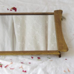 May include: A small, rectangular mirror with a gold-colored frame. The frame has a simple, elegant design. The mirror is resting on a white surface with floral details and scattered red petals.