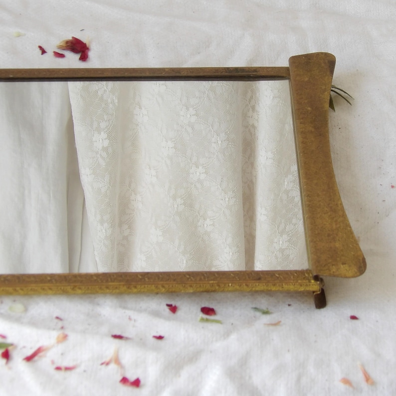 May include: A small, rectangular mirror with a gold-colored frame. The frame has a simple, elegant design. The mirror is resting on a white surface with floral details and scattered red petals.