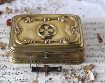 May include: Antique brass warming tray with a decorative design and a handle. The tray has a circular opening with a metal grill inside. The tray is sitting on a sheet of music.