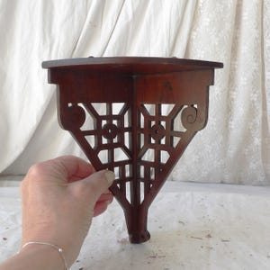 May include: A dark brown wooden corner shelf featuring a flat triangular top surface and an intricate, openwork support bracket with geometric square patterns and decorative scroll accents along the sides, designed for wall mounting.