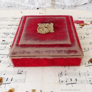 May include: A red velvet jewellery box with a gold-toned clasp decorated with clear rhinestones. The box is closed and sitting on a sheet of music.