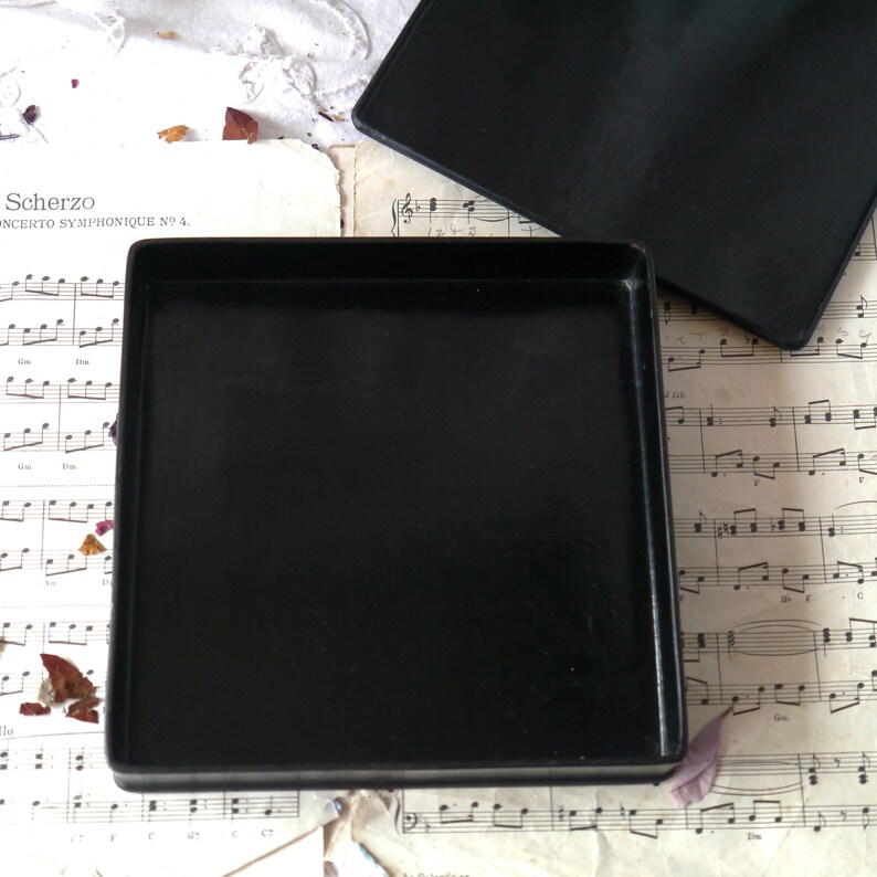 Pu&ograve; includere: Una scatola quadrata nera con un coperchio. La scatola &egrave; vuota e il coperchio &egrave; aperto. La scatola &egrave; appoggiata su un foglio di musica.