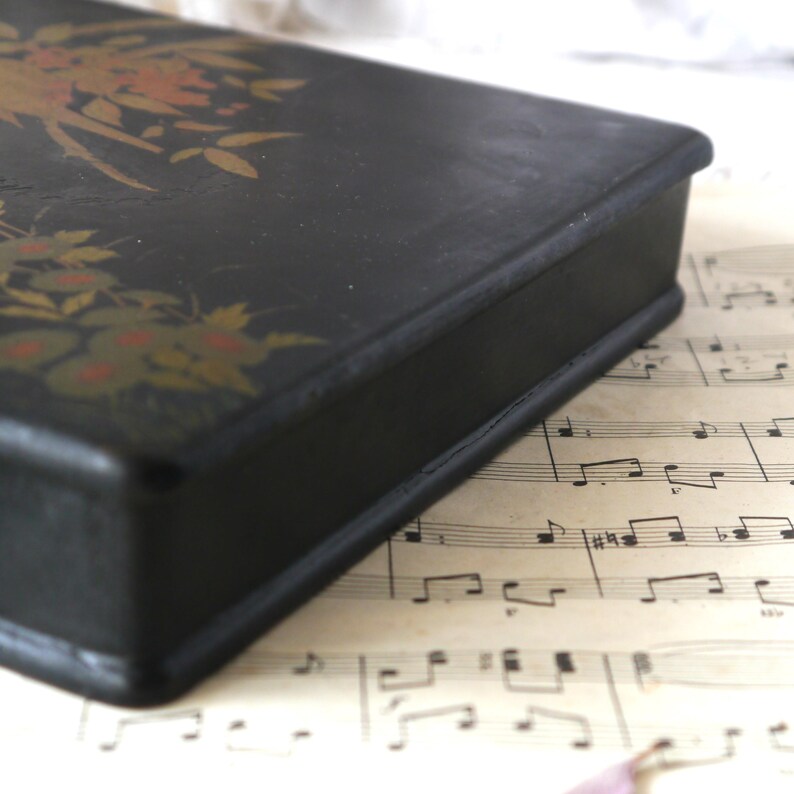 Pu&ograve; includere: Una scatola di legno nera con un disegno floreale dipinto sulla parte superiore. La scatola &egrave; chiusa e si trova su un foglio di musica.