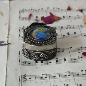 以下が含まれることがあります： 装飾的なデザインとカラフルなモザイクガラスの蓋が付いた銀製の小物入れ。音楽の楽譜の上に置かれています。