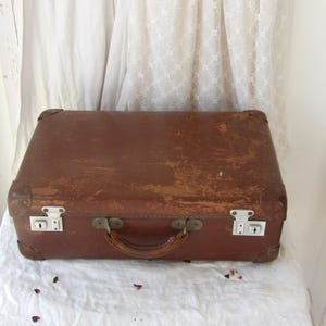 May include: A well-worn brown suitcase, showing signs of age and use. It features metal corner reinforcements, two metal clasps, and a leather handle. The suitcase is placed on a white surface.