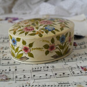 May include: A white oval trinket box with a floral design. The box is painted with pink, blue, yellow, and green flowers. The box is sitting on a sheet of music.