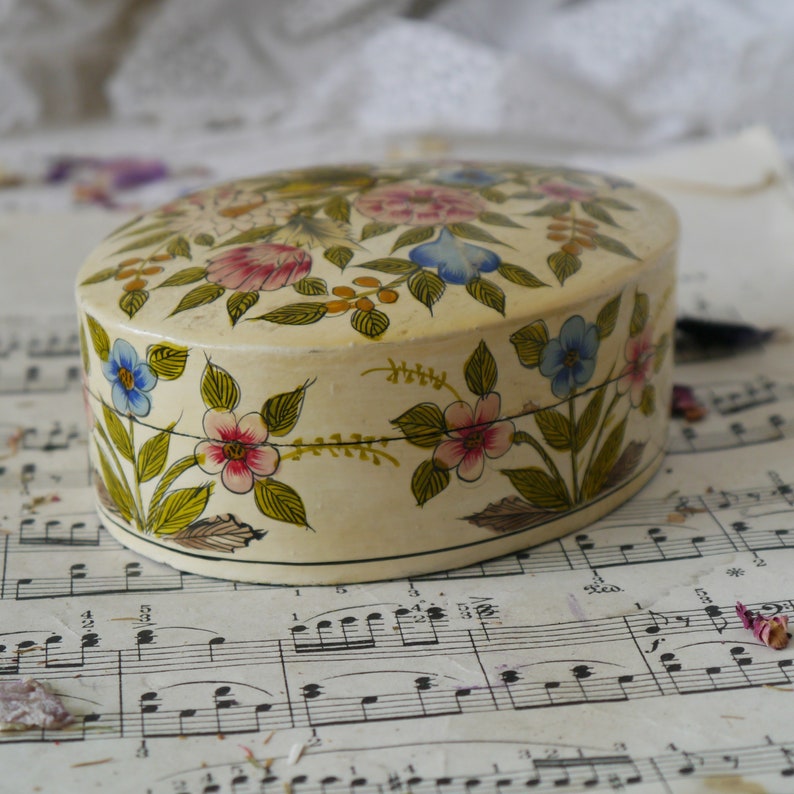 May include: A white oval trinket box with a floral design. The box is painted with pink, blue, yellow, and green flowers. The box is sitting on a sheet of music.