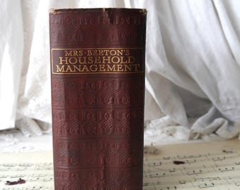 Op de afbeelding: Een vintage, rood, in leer gebonden boek getiteld "Mrs. Beeton's Household Management" met gouden letters. Het boek ligt op een blad muziek met rode bloemblaadjes eromheen.