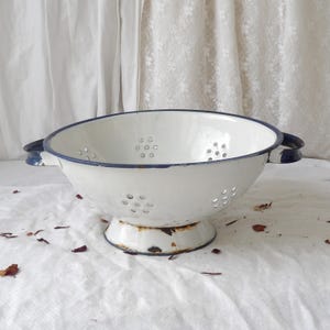 Pode incluir: Escorredor de esmalte branco com borda e alças azuis. O utensílio de cozinha vintage tem uma base de pedestal e inúmeros orifícios de drenagem. O escorredor mostra sinais de desgaste, adicionando ao seu charme antigo. Uma ferramenta de cozinha clássica.