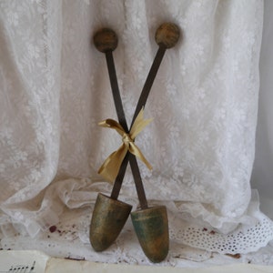 May include: A pair of wooden shoe trees with a gold ribbon tied around the handles. The shoe trees are crossed and the handles are pointing up. The shoe trees are on a white surface with a lace tablecloth.