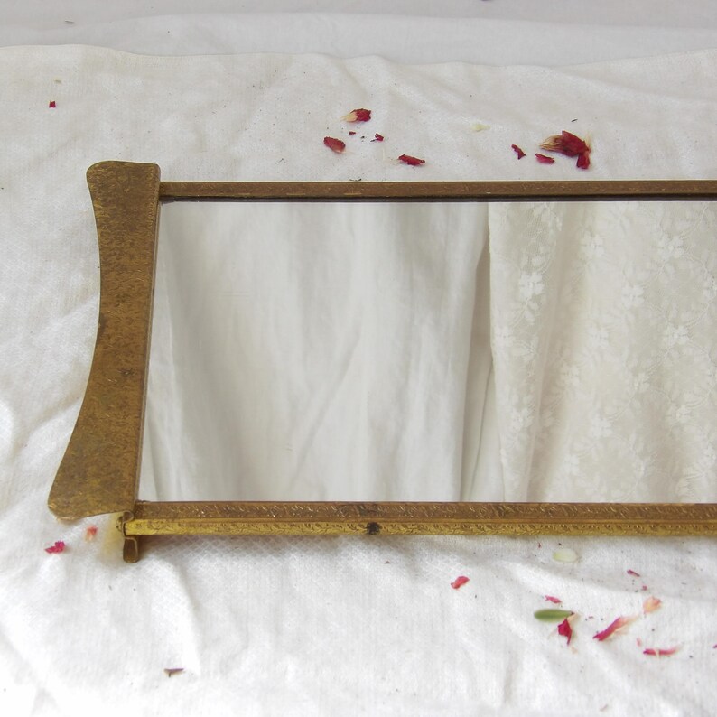 May include: A rectangular, antique-style mirror with a gold-colored frame. The frame has a decorative, textured design and a curved support on one side. The mirror is set against a white background with scattered rose petals.