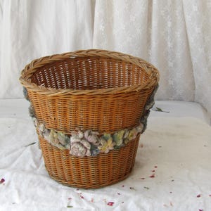 Può includere: Un cesto rotondo in vimini intrecciato di colore marrone caldo, adornato con una fascia decorativa di fiori scolpiti. La fascia floreale presenta fiori rosa, verdi e color crema. Il cesto è probabilmente utilizzato per la conservazione o l'esposizione.