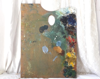 May include: A rectangular wooden artist palette featuring a thumb hole at the top center. The surface is covered in dried paint splatters and thick, textured dabs of various colors including blue, green, yellow, red, brown, and black.