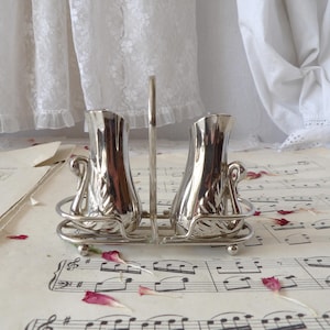May include: A vintage silver salt and pepper shaker set. The set includes two ornate shakers with a decorative design, held in a matching metal stand with a handle. The set is displayed on a sheet of vintage sheet music, with scattered red flower petals.