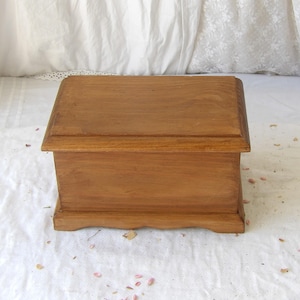 May include: A rectangular wooden box with a medium brown finish, featuring a flat lid with beveled edges and a base with a scalloped bottom edge. The box has a smooth, consistent wood grain texture and a simple, functional design.