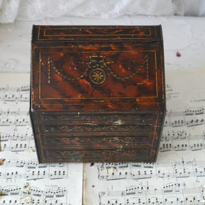 May include: A vintage, small, decorative box with a hinged lid and three small drawers. The box is primarily brown with a tortoiseshell pattern and gold-colored accents. The front features a gold floral design and the drawers have gold scrollwork.