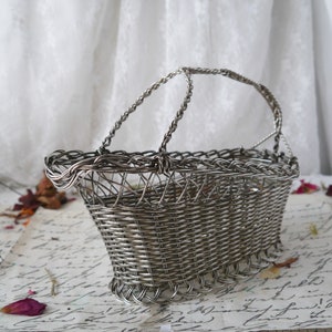 May include: A silver wire basket with a woven design and two handles. The basket is sitting on a piece of paper with writing on it.