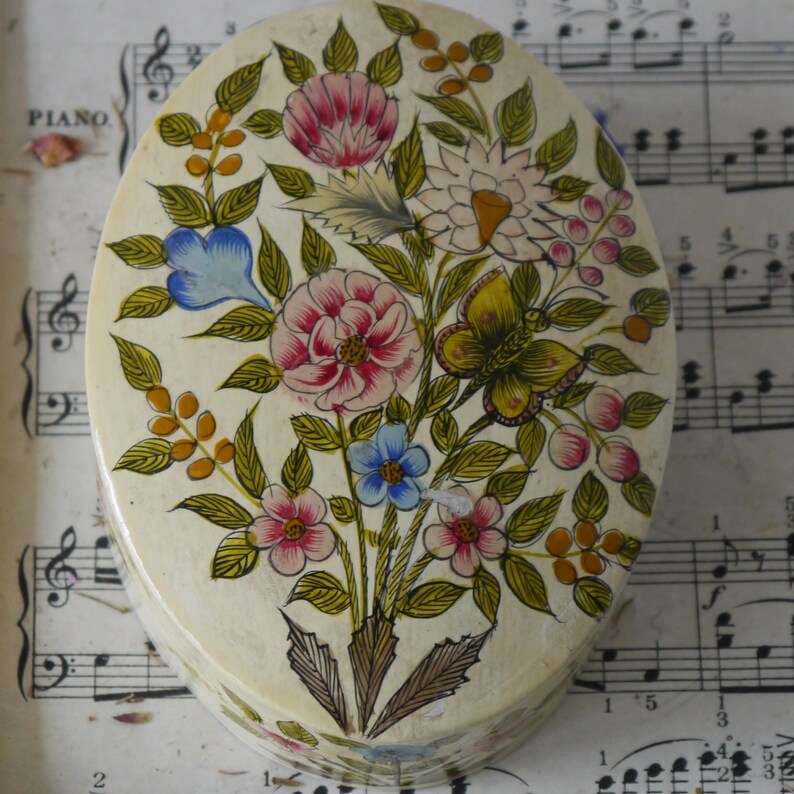 May include: An oval-shaped trinket box with a white background and a floral design painted in shades of pink, blue, yellow, and green. The flowers are stylized and have a whimsical feel. The box is perfect for storing small treasures or trinkets.