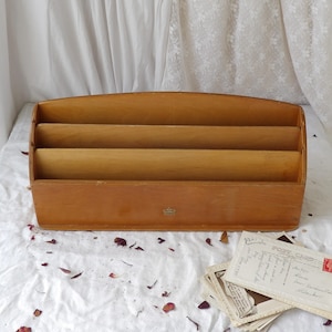 May include: A vintage wooden letter rack with three compartments. The organiser is made of light brown wood and has a curved top. A small emblem is visible on the front. Postcards and dried petals are scattered around the organiser.