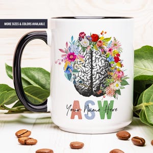 May include: White ceramic mug with a black rim and handle. The mug features a colorful floral design surrounding a black and white illustration of a human brain. The text "Your Name Here" is printed below the design in rainbow colors.