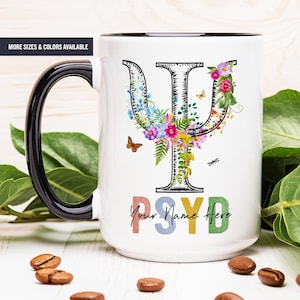 May include: White ceramic mug with a black rim and handle. The mug features a floral design around a black and white Greek letter psi symbol. The text "Your Name Here" is written in rainbow colors below the symbol.