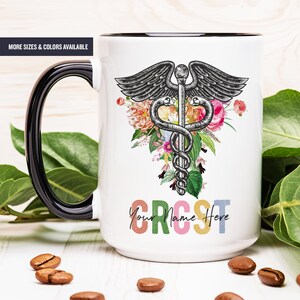 May include: White ceramic mug with a black rim and handle. The mug features a black and white medical caduceus symbol with a floral design. The text "Your Name Here" is printed in rainbow colors at the bottom of the mug.