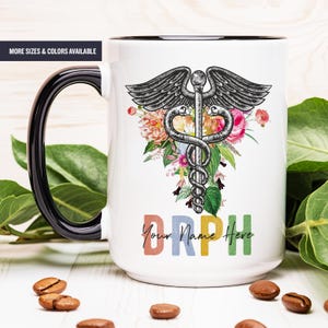 May include: White ceramic mug with a black rim and handle. The mug features a black and white medical caduceus symbol with a floral design and the text "DRPH Your Name Here" in rainbow colors.