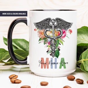 May include: White ceramic mug with a black rim and handle. The mug features a black and white medical caduceus symbol with a floral design. The text "Your Name Here MHA" is printed on the mug in colorful letters.