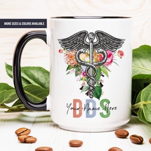May include: White ceramic mug with a black handle and rim, featuring a Caduceus symbol with floral accents. The mug has the text "DDS Your Name Here" in colorful letters. The words "MORE SIZES & COLORS AVAILABLE" are also visible.