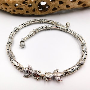 May include: Silver necklace with a detailed design featuring two fish charms. The necklace is made of small, round beads with intricate patterns.