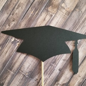 May include: Black graduation cap cutout on a stick. The cap has a tassel. The cutout is on a wood background.
