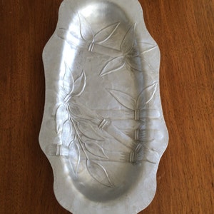 May include: Silver-colored, oblong serving tray with a scalloped edge. The tray features an embossed bamboo and leaf design. The tray is made of metal and sits on a wooden surface.