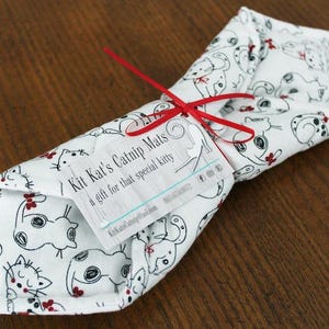 May include: A white catnip mat with a black and white cat print. The mat is rolled up and tied with a red ribbon. A white tag is attached to the mat with the text "Kit Kat's Catnip Mats a gift for that special kitty".