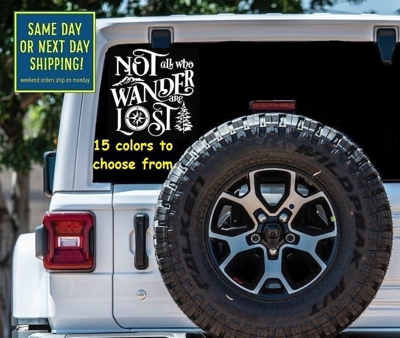 May include: White vinyl decal with the text "NOT all who WANDER are LOST" featuring a compass and pine tree graphic. The decal is on the back of a white Jeep. The text "15 colours to choose from" is below the decal.