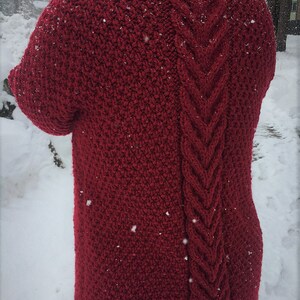 May include: A red knitted sweater with a cable knit pattern on the back. The sweater is photographed in a snowy setting.