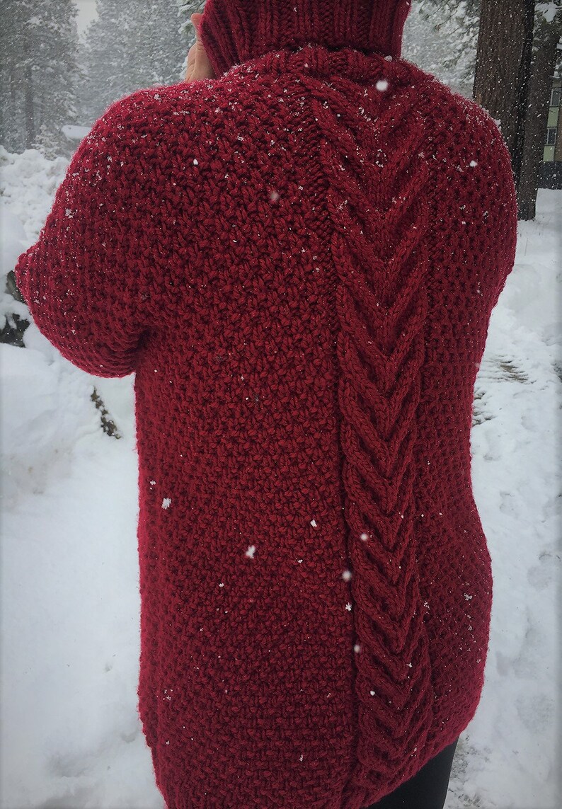 May include: A red knitted sweater with a cable knit pattern on the back. The sweater is photographed in a snowy setting.