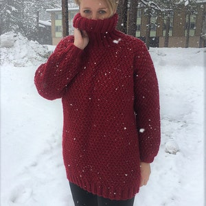May include: A red knitted turtleneck sweater with a textured pattern. The sweater is worn by a person standing in the snow.
