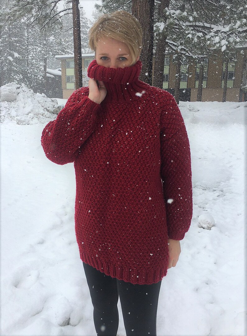 May include: A red knitted turtleneck sweater with a textured pattern. The sweater is worn by a person standing in the snow.