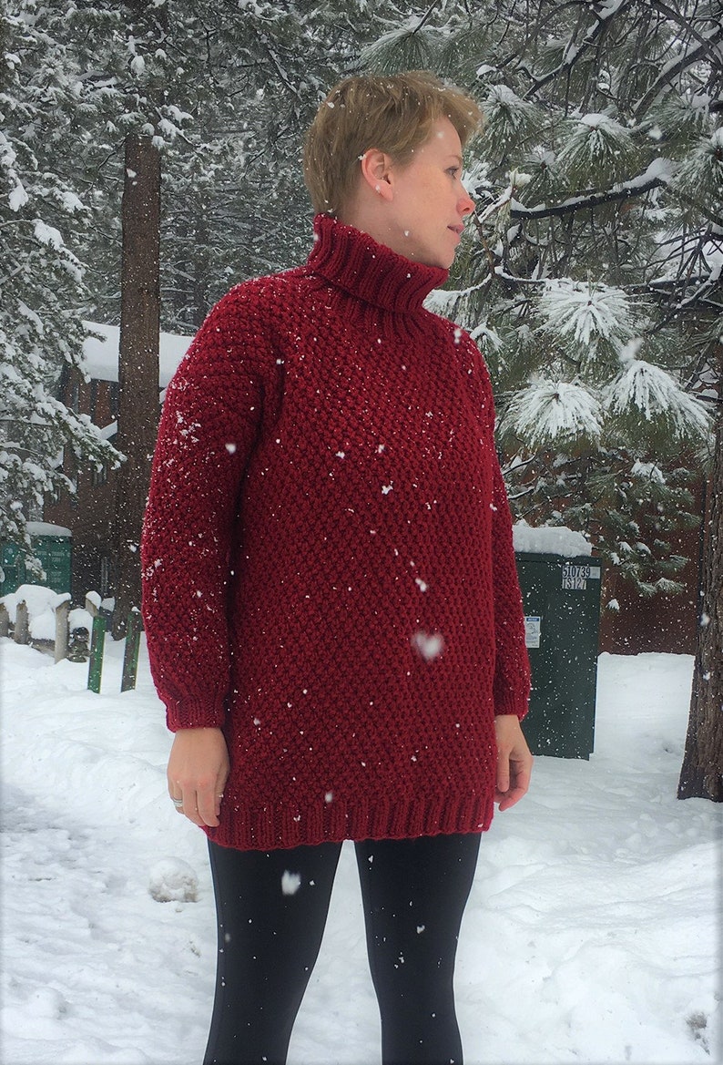 May include: A red knitted turtleneck sweater with a textured pattern. The sweater is worn by a person standing in a snowy landscape.