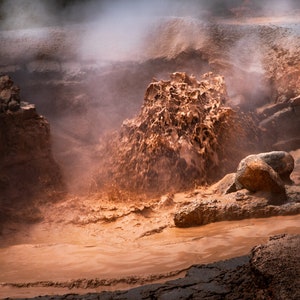 May include: A close-up of a mud volcano erupting, with steam rising from the crater. The mud is a rich brown color and has a textured surface. The surrounding area is rocky and barren.
