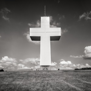 May include: A large white cross stands tall against a cloudy sky. The cross is made of concrete and has a simple design. It is located on a grassy hilltop.
